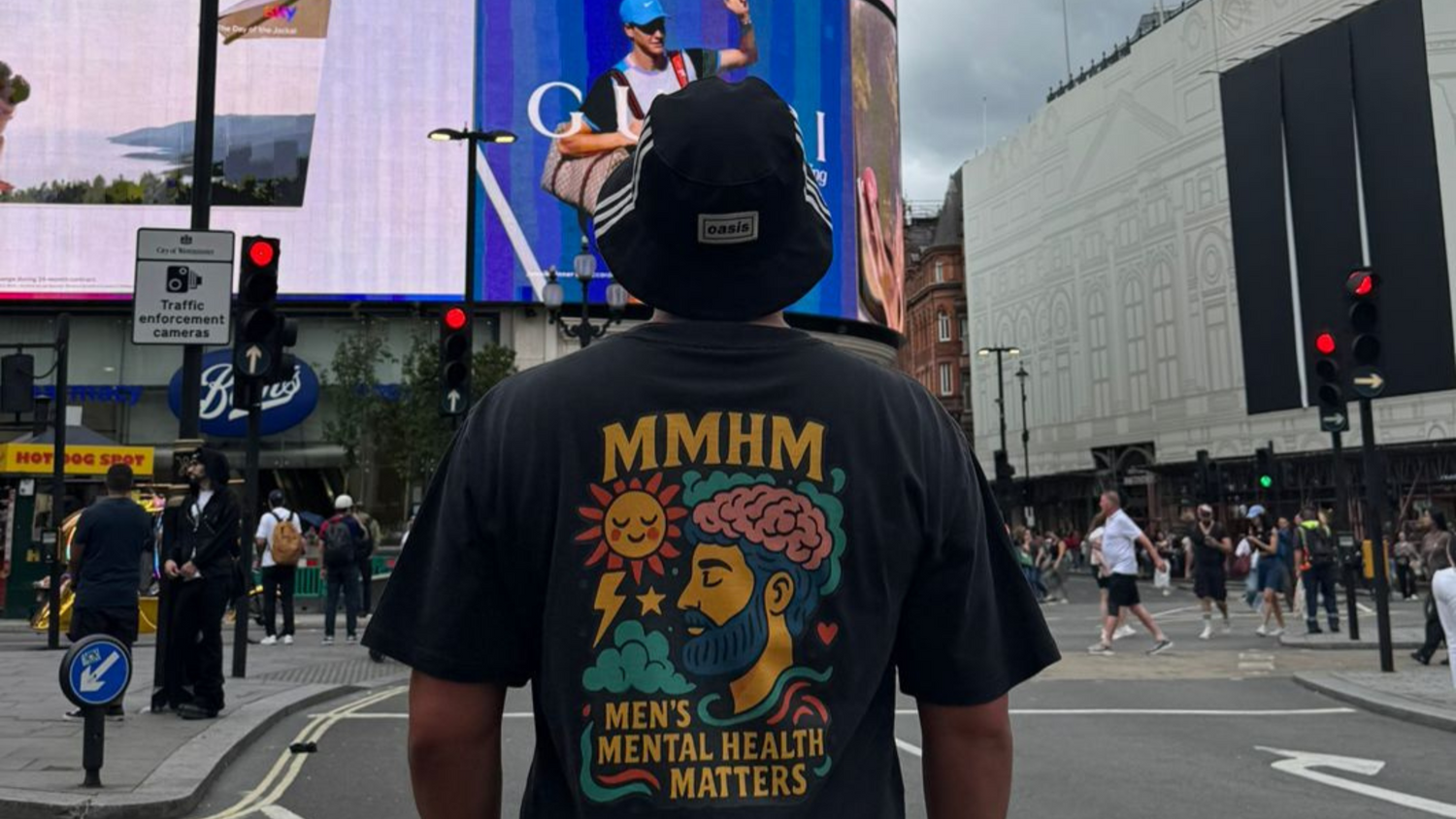 Person wearing a black t-shirt with 'Mens Mental Health Matters' on a busy street with a large digital billboard in the background.